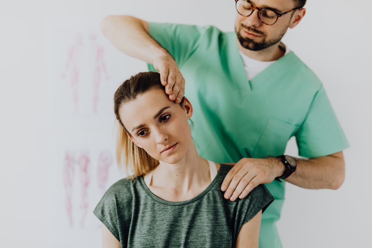 services-01 Crop osteopath in uniform and eyeglasses standing with closed eyes while examining neck of young thoughtful female in casual wear in hospital